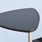 Close-up of a modern, triangular black tabletop with rounded corners, supported by a single metal leg. The surface appears glossy, and the background is a plain, light-colored wall.