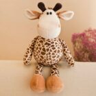 A plush giraffe toy with large eyes and a patterned body sits upright on a light-colored surface. Its hooves are brown, and it has small horns. In the background, a pink plant decorates the beige wall.
