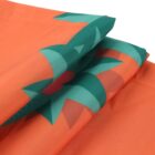 Close-up of folded fabric in a bright orange hue, featuring a bold geometric design in green and red. The design appears abstract, with sharp angles and symmetrical shapes, adding a dynamic contrast against the solid orange background.