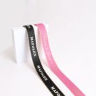 Two satin ribbons draped over a white cube. The black ribbon features the word MAFOLEN in white, while the pink ribbon has delicate cursive text in gold. The background is a smooth, light gray.