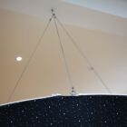 Close-up of a black object with a star pattern, suspended from the ceiling by metal cables. The background shows a ceiling with recessed lighting and a small spot of illumination.