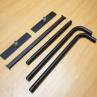 Disassembled black metal handrails and mounts are laid out on a wooden floor. The components include two straight metal rods, two curved rods, and two rectangular mounting plates with pegs.