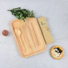A wooden tray with a spoon, fork, chopsticks, and straw on it. A rolled mat is next to the tray on a textured surface. A bowl with black and white beans and a sprig of leafy greens are also present.