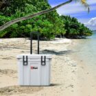 A white cooler with black handles and a logo stands on a sandy beach near clear blue water. Lush green trees and a palm tree with a curved trunk line the background. The sky is partly cloudy and sunny.