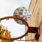 A colorful, floral-patterned basketball is caught mid-air above a rusty hoop mounted on a stone wall. The sky is overcast, and a tree with autumn leaves is visible in the background.