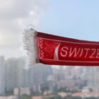 A red scarf with white fringes hangs near a window, displaying a design and the partial word SWITZ. The view outside shows a cityscape with tall buildings under a partly cloudy sky.