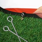 Artificial turf with two metal stakes and a corner showing orange and beige fabric. The stakes have hooked ends, and the setup suggests a camping or outdoor theme.