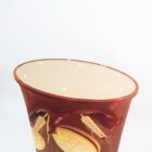 Oval-shaped container with a design featuring wheat stalks and crackers against a chocolate brown background. The top is open with evenly spaced holes, and the container’s design is vivid against the white backdrop.