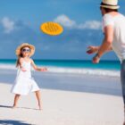A child in a white dress and straw hat plays frisbee with an adult wearing a hat and sunglasses on a sandy beach, with the ocean and blue sky in the background.