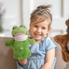 A young girl with a tiara smiles while sitting on a couch, holding a green frog puppet. Shes wearing a light blue dress, and theres another puppet partially visible on the right. The background is softly blurred.