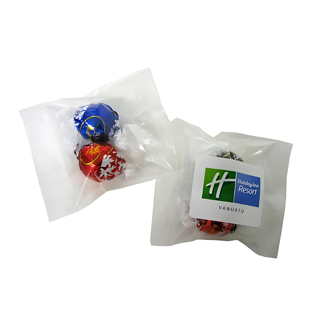 Two small clear cello bags, each with 2 Lindt Balls inside, are wrapped in colorful foil with white snowflake designs. One bag bears a label featuring the green and white Holiday Inn Resort Vanuatu logo.