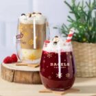 Two festive drinks in transparent mugs rest on wooden slabs. The front mug holds a red drink with berries and a candy cane straw, labeled BAGELS FACTORY. The back mug contains a frothy coffee labeled Arab 1965. A small pine tree is in the background.