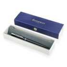 A sleek black pen with silver accents rests in an open cream-colored box lined with gray padding. The box lid, displaying the Waterman Paris logo, is positioned behind it, featuring a blue design.