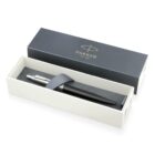 A Parker pen placed diagonally inside an open white box with a gray lid. A gray elastic band secures the pen. The lid, positioned to the side, displays the Parker logo with EST. 1888 in gold.