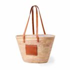 A woven straw tote bag with long brown leather handles and a square leather patch on the front. The bag has an open top and a simple, rustic design.