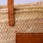 Close-up of a woven straw basket with a leather handle and stitched leather pocket. The basket features an intricate pattern, and the leather elements are a rich brown color, providing a contrast to the natural straw texture.