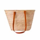 A woven straw basket with two long brown leather handles, set against a plain white background.