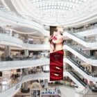 A large indoor shopping mall with multiple escalators and storefronts. A vertical banner featuring a womans face with red lipstick and two lipstick tubes is prominently displayed in the center. The mall is well-lit with a glass ceiling.