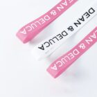 Three fabric straps on a white surface, two are pink, and one is white, each with the text DEAN & DELUCA in bold white or black letters. The straps are arranged diagonally, with one pink strap slightly ahead of the others.