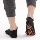Two feet wearing black non-slip socks with orange grip patterns on the soles. The person is wearing gray pants, and the image shows a back view of the feet on a white background.