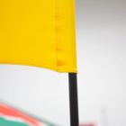 Close-up of a bright yellow flag on a black pole, partially cropped. In the background, blurred stripes of red, white, and green suggest other flags or decorations.