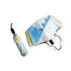 A compact, folded umbrella with a geometric pattern of blue, white, and yellow sits next to a matching tote bag. The umbrella handle has a wrist strap. Both items feature abstract triangles and circular shapes on a white background.