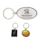 Three keychains: an oval one with a Peugeot logo, a rectangular one with Gary Greer text on a dark background, and a round one with a striped orange, yellow, and red pattern.