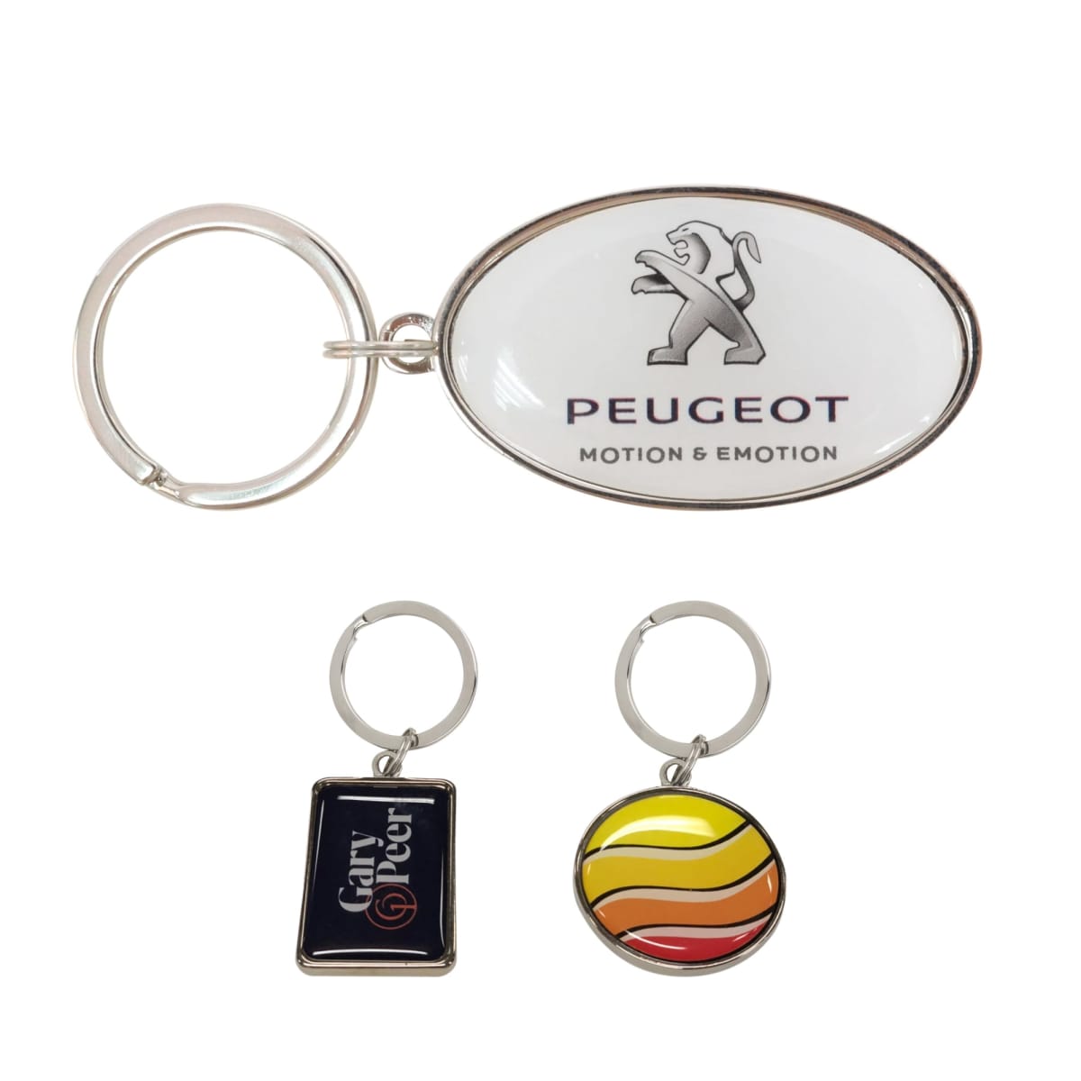 Three keychains: an oval one with a Peugeot logo, a rectangular one with Gary Greer text on a dark background, and a round one with a striped orange, yellow, and red pattern.