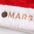 Close-up of a red and white Santa hat with brown embroidered text MARS and a globe-like design to the left on the white brim.