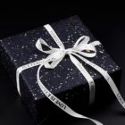 A gift box wrapped in dark speckled paper tied with a white ribbon that says LOVE IS ETERNAL in black text. The box is placed on a black surface.