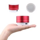 A hand holding a small, cylindrical red and white speaker with control buttons and a USB port. A top view of the speaker is shown above, displaying its mesh speaker grille. The speaker has a compact and portable design.