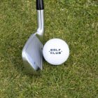 A close-up of a golf iron next to a golf ball on green grass. The golf ball has GOLF CLUB printed on it in black text.