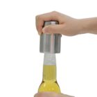 A hand using a stainless steel bottle opener to remove the cap from a glass bottle filled with a yellow liquid. The background is plain white.