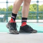 Close-up of a person wearing black sneakers and colorful socks with basketball designs, standing on a sports court. The background shows a fenced area and greenery.