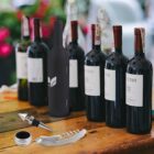 Seven wine bottles, one with a logo, are lined up on a wooden table outdoors. In front of them, there is a wine stopper, a drip ring, and a corkscrew. The background features blurred greenery and flowers.