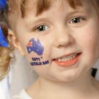 Smiling child with light brown hair and blue ribbon, with temporary tattoo of Australia and flag on cheek reading Happy Australia Day.