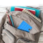 A beige backpack is open, revealing notebooks, a spiral-bound book, pencils, and a blue pop-it toy inside its main compartment. The backpack rests on a white wooden surface.
