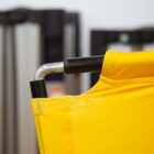 Close-up of the corner of a yellow fabric chair or cot with a metal frame, featuring an angular joint. The background is blurred, showing some vertical objects, possibly equipment or furniture, in shades of gray and black.