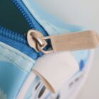 Close-up of a blue zipper on a light blue fabric with a metallic zipper pull. The fabric has subtle white and darker blue patterns.