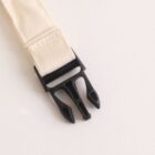 Close-up of a beige fabric strap with a black plastic buckle on a light surface. The buckle is open, showing two prongs and slots for fastening. The stitching on the strap is visible, emphasizing its texture and construction.