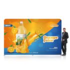 A man in a suit stands next to a vibrant orange and blue sign featuring a large bottle of orange juice and splashing juice. The sign includes sliced oranges and text that reads Orange Juice with New! and 100% Natural Ingredients.
