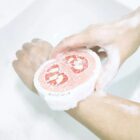 A person washing their hands with a round soap that resembles a grapefruit slice, creating a lather over a sink. The soap has pink and white colors, mimicking the fruits appearance.