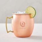 Copper mug with ice and a lime slice on the rim, featuring a brass handle. A company logo is printed on the mug. The background is plain and neutral.
