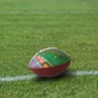 A football with an image of a stadium and crowd printed on its surface rests on a green, grassy field near a white line. The field is well-maintained, with vibrant, lush grass.