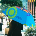 A person holds a colorful umbrella with green circles and blue sections, and the word ADIO printed on it. They are walking down a city street lined with greenery and buildings in the background.