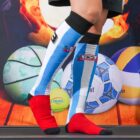 Person wearing knee-high sports socks with red feet and blue, white, and black stripes stands in front of a colorful backdrop featuring basketballs, volleyballs, and a soccer ball.