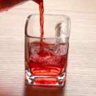 Red liquid being poured into a square glass on a light wooden surface. The liquid has bubbles as it fills the glass, creating an effervescent effect. The word ADIO is visible on the side of the glass.