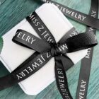 A small white gift box tied with a black ribbon that reads MISS Z JEWELRY in white letters, placed on a textured teal surface. The ribbon forms a neat bow on top of the box.