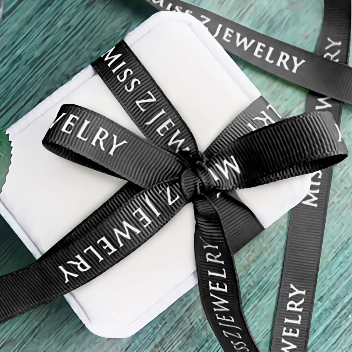 A small white gift box tied with a black ribbon that reads MISS Z JEWELRY in white letters, placed on a textured teal surface. The ribbon forms a neat bow on top of the box.