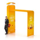 Man in a suit leans casually against a bright, orange structure with World Emoji Day text. The structure is adorned with colorful, smiling emojis and has a rooftop element.